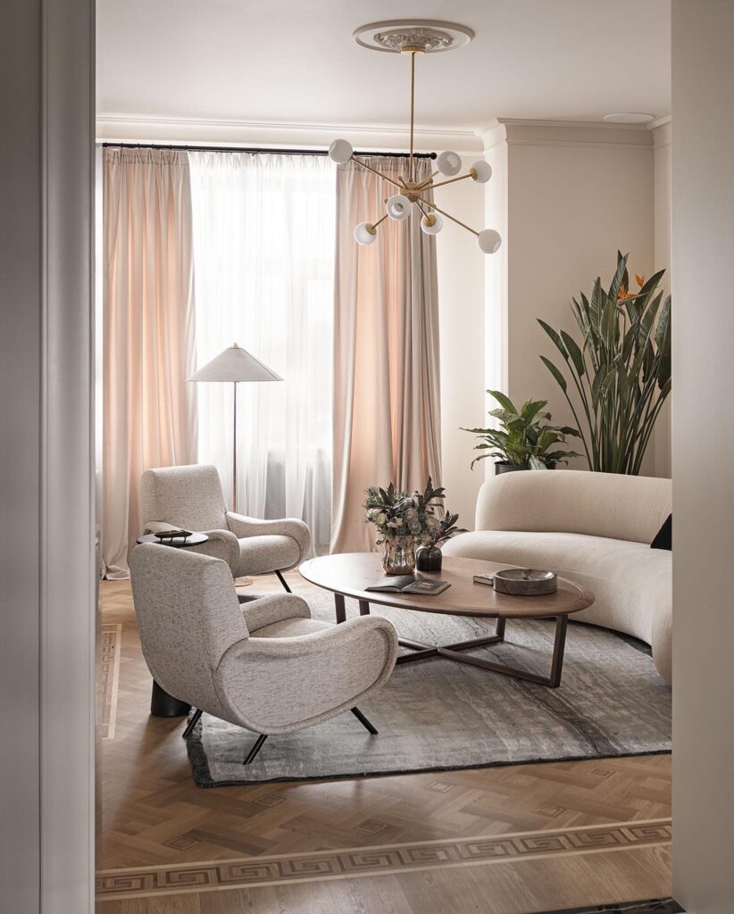 Elegant living room design by Sivak+Partners featuring a modern chandelier, plush gray furniture, and lush potted plants in a warm, neutral color palette with high-end finishes.