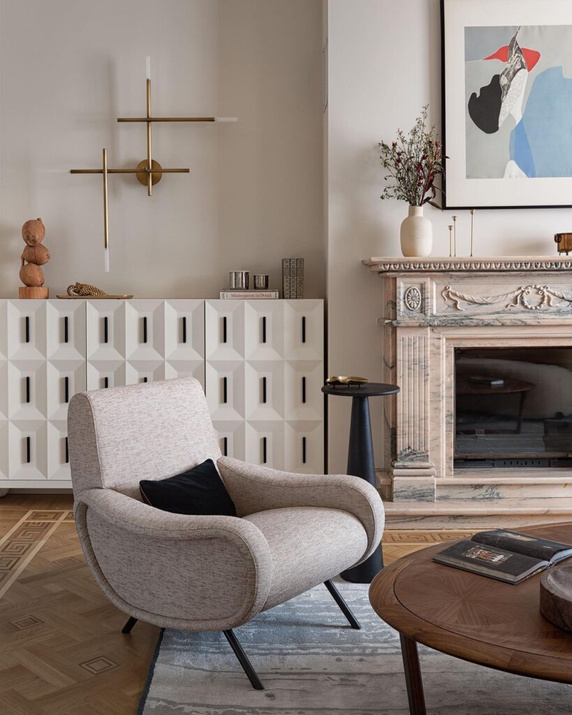 A cozy living room with a modern, minimalist design featuring a grey upholstered armchair, a wooden coffee table, and a decorative fireplace mantel. The room also includes abstract artwork, a vase of flowers, and built-in storage cabinets with a geometric pattern.