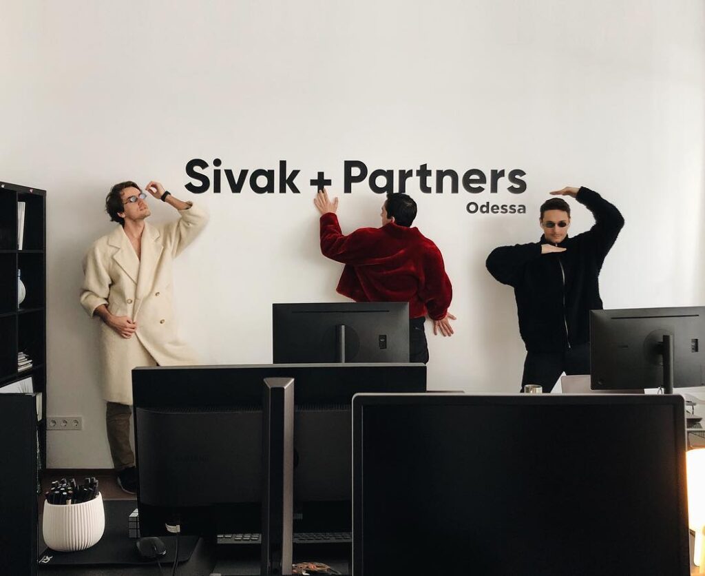 Employees Posing In Front Of Sivak + Partners Design Studio Logo