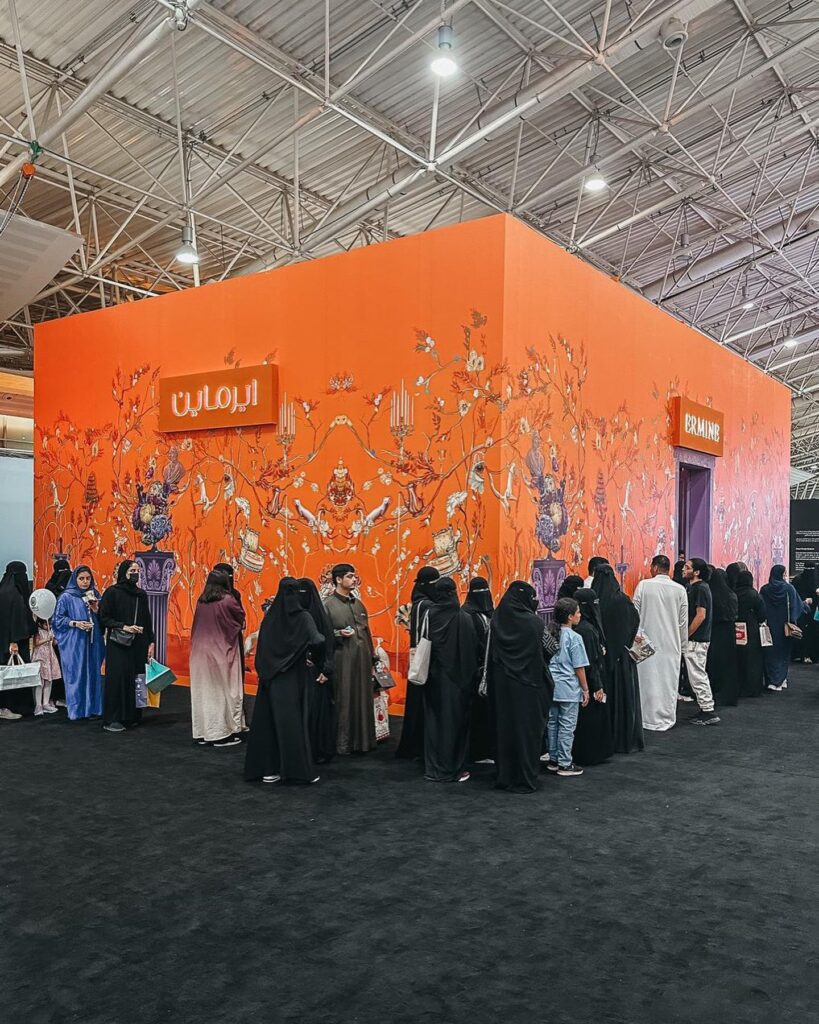 Alt Text: Vibrant orange wall with intricate floral and abstract patterns serves as backdrop for group of people in exhibition or event space. Arabic word "Aramain" prominently displayed, suggesting design or architectural theme. People appear to be visitors or attendees, some wearing traditional Middle Eastern attire. High-ceilinged space with metal truss structure indicates commercial or industrial setting, likely design or art exhibition.