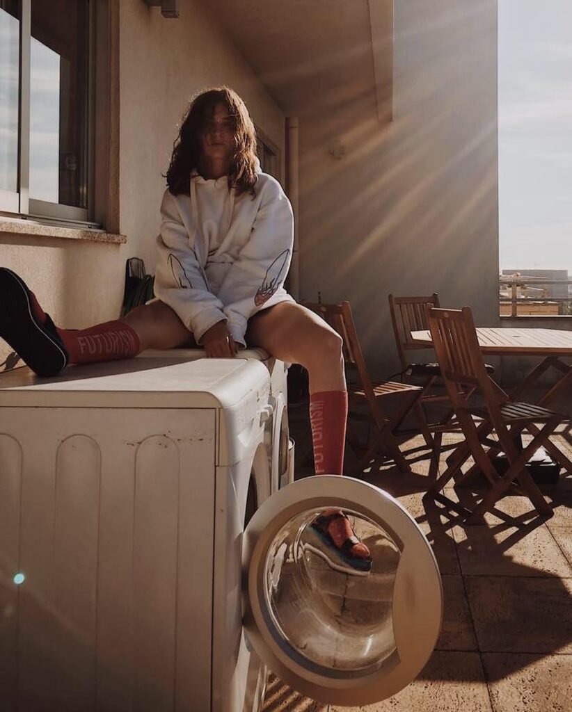 A Woman Sitting On A Washing Machine In A Cozy Indoor Setting With Wooden Furniture And Natural Lighting